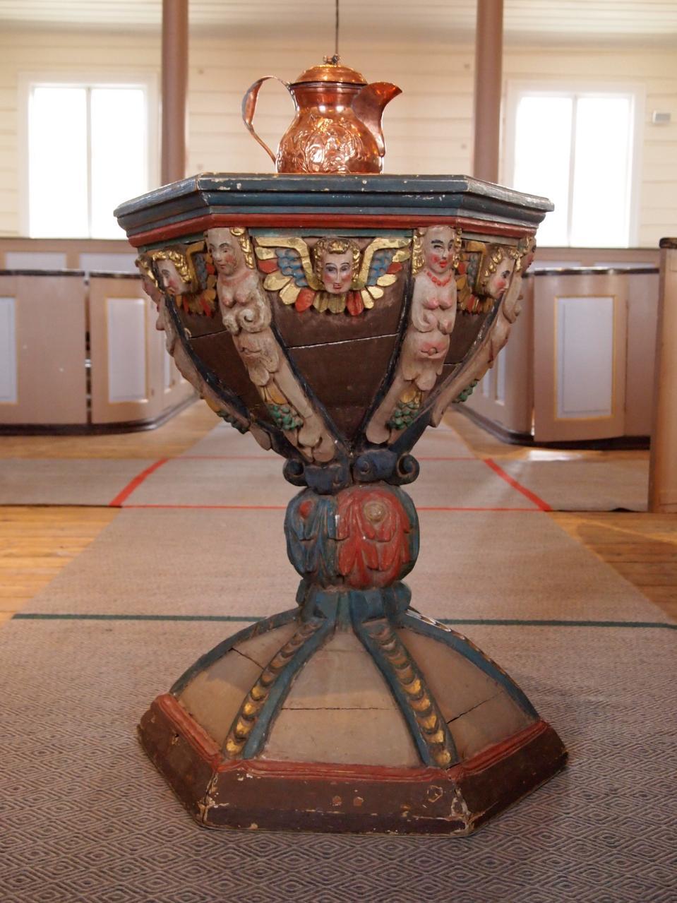 DØPEFONT Foran alteret står døpefonten. Den er sekskantet, timeglassformet, og har utskårne figurer og ornamenter.